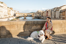 Stylish Woman Walks With A Dog On A Bridge In Florence, Visiting Italian Landmarks And Traveling With Italian Shepherd Dog Maremma