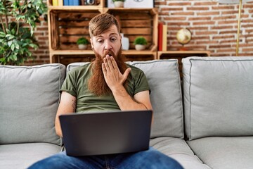 Sticker - Redhead man with long beard using laptop sitting on the sofa at the living room covering mouth with hand, shocked and afraid for mistake. surprised expression