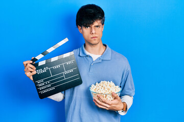 Poster - Handsome hipster young man eating popcorn holding cinema clapboard skeptic and nervous, frowning upset because of problem. negative person.