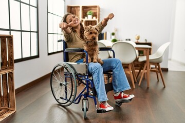 Wall Mural - Young hispanic girl sitting on wheelchair at home smiling and confident gesturing with hand doing small size sign with fingers looking and the camera. measure concept.