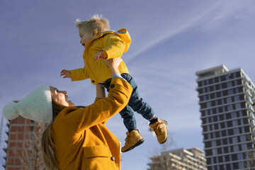 A cute little boy has fun with his mother on a sunny day in the town. They are both wearing yellow jackets, and mom throws him up in the air, which causes a big smile on the boy's face.