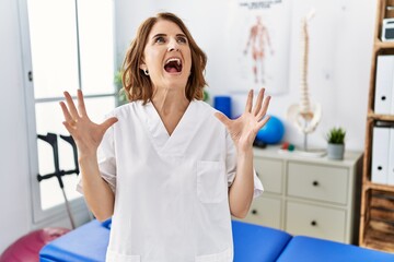 Canvas Print - Middle age physiotherapist woman working at pain recovery clinic crazy and mad shouting and yelling with aggressive expression and arms raised. frustration concept.