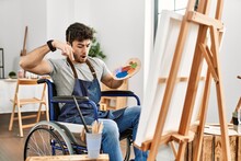 Young Hispanic Man Sitting On Wheelchair Painting At Art Studio Pointing Down With Fingers Showing Advertisement, Surprised Face And Open Mouth