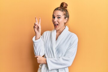 Canvas Print - Young blonde woman wearing bathrobe smiling with happy face winking at the camera doing victory sign. number two.