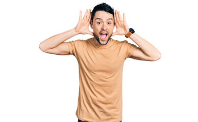 Hispanic man with beard wearing casual t shirt smiling cheerful playing peek a boo with hands showing face. surprised and exited