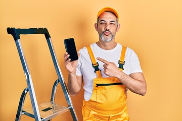 Canvas Print - Handsome middle age man with grey hair standing by ladder showing smartphone looking at the camera blowing a kiss being lovely and sexy. love expression.