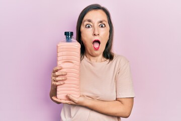 Poster - Middle age hispanic woman holding detergent bottle afraid and shocked with surprise and amazed expression, fear and excited face.