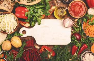 Wall Mural - Various uncooked vegetables, herbs and spices for healthy cooking and white cutting board on rustic kitchen wooden table, top view. Preparation vegetarian cabbage, beetroot, tomato soup. 