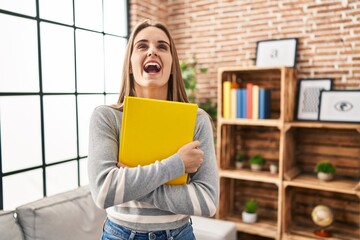 Wall Mural - Young woman holding notebook angry and mad screaming frustrated and furious, shouting with anger looking up.