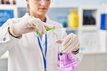 Poster - Young blonde woman wearing scientist uniform pouring liquid on test tube at laboratory