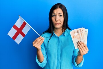 Sticker - Young latin woman holding england flag and pounds banknotes skeptic and nervous, frowning upset because of problem. negative person.