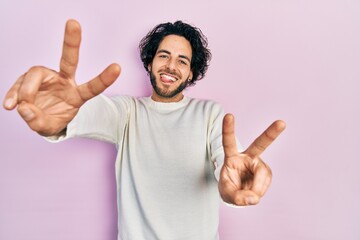 Wall Mural - Handsome hispanic man wearing casual white sweater smiling with tongue out showing fingers of both hands doing victory sign. number two.