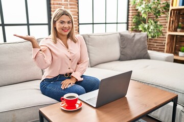 Poster - Young hispanic woman using laptop sitting on the sofa at home smiling cheerful presenting and pointing with palm of hand looking at the camera.