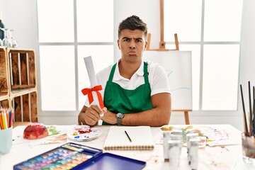 Sticker - Young hispanic man at art studio holding degree depressed and worry for distress, crying angry and afraid. sad expression.