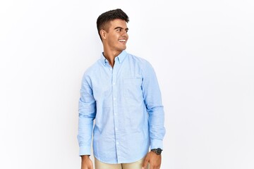 Young hispanic man wearing business shirt standing over isolated background looking away to side with smile on face, natural expression. laughing confident.