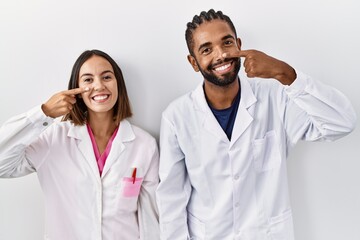 Wall Mural - Young hispanic doctors standing over white background pointing with hand finger to face and nose, smiling cheerful. beauty concept