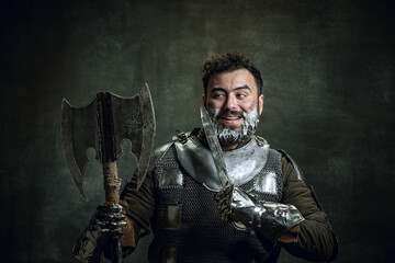 Comic portrait of medieval warrior or knight with dirty wounded face shaving with an ax isolated over dark vintage background. Comparison of eras, history, beauty