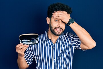 Poster - Young arab man with beard holding glasses in eyewear case stressed and frustrated with hand on head, surprised and angry face