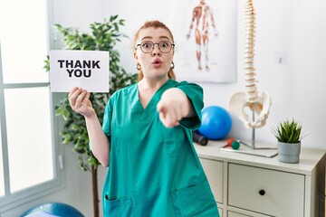 Poster - Young redhead physiotherapist woman working at pain recovery clinic holding thank you banner pointing with finger to the camera and to you, confident gesture looking serious