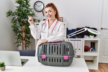 Canvas Print - Young redhead veterinarian woman working at pet clinic smiling happy pointing with hand and finger to the side