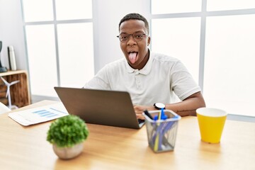 Poster - Young african man working at the office using computer laptop sticking tongue out happy with funny expression. emotion concept.