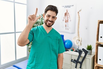 Poster - Young man with beard working at pain recovery clinic showing and pointing up with fingers number two while smiling confident and happy.