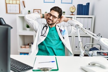 Poster - Young man with beard wearing doctor uniform and stethoscope at the clinic relaxing and stretching, arms and hands behind head and neck smiling happy