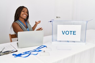 Wall Mural - Young african american woman working at political election sitting by ballot inviting to enter smiling natural with open hand