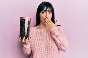 Canvas Print - Young hispanic woman holding jar with coffee beans covering mouth with hand, shocked and afraid for mistake. surprised expression