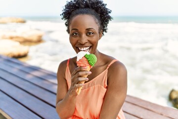 Poster - Young african american woman smiling happy on a summer day by the beach eating a ice cream