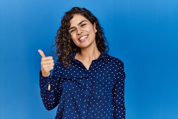 Poster - Young brunette woman with curly hair wearing casual clothes over blue background doing happy thumbs up gesture with hand. approving expression looking at the camera showing success.