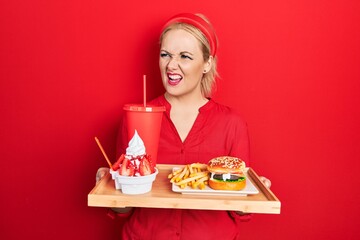 Canvas Print - Young blonde woman eating a tasty classic burger with fries and soda angry and mad screaming frustrated and furious, shouting with anger. rage and aggressive concept.