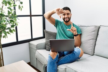 Canvas Print - Young handsome man with beard using computer laptop sitting on the sofa at home smiling making frame with hands and fingers with happy face. creativity and photography concept.