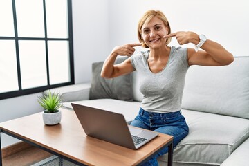 Sticker - Middle age blonde woman using laptop at home smiling cheerful showing and pointing with fingers teeth and mouth. dental health concept.