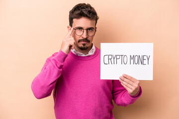 Young caucasian man holding a crypto money placard isolated on beige background pointing temple with finger, thinking, focused on a task.