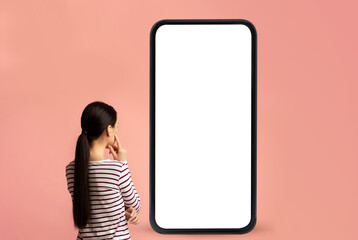 Wall Mural - Back view of woman looking at big smartphone with blank white screen and thinking, device with space for mockup