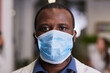 Young serious African-American healthcare worker in protective mask and workwear standing against hospital interior and looking at camera