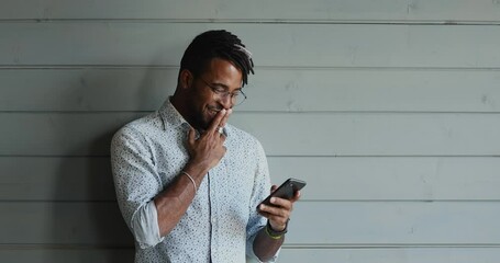 Handsome stylish young Afro American man in glasses stand holding mobile phone use messenger for web dating chatting online read email create answer with smile. Grey fence is on background