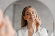 © Prostock-studio - Young woman caring for facial skin using cotton pad and looking in round mirror in bathroom interior