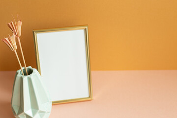 Wall Mural - Blank photo frame with vase of dry flowers on pink table. orange wall background. copy space