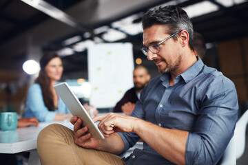 Poster - The creative process. Shot of a man sitting at a table in an office using a digital tablet with colleagues working in the background.