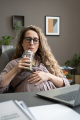 Wall Mural - Young pregnant businesswoman drinking fresh cocktail while sitting at her workplace in front of the laptop at office