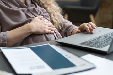 Wall Mural - Close-up of pregnant businesswoman touching her belly while sitting at the table and typing on laptop during online work at office