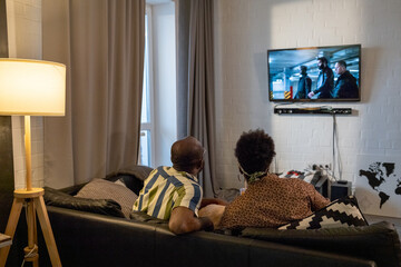 Wall Mural - Rear view of African married couple watching TV together hanging on the wall while resting on sofa in the living room