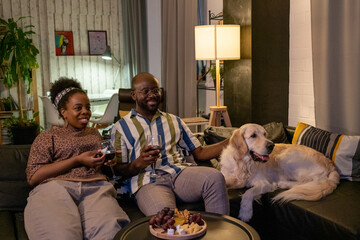 Wall Mural - Happy African couple drinking wine and watching movie while resting on sofa with their dog, they spending time at home in the evening