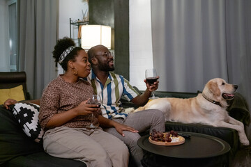 Wall Mural - Happy African couple drinking red wine and eating fruits while resting on sofa together with dog and watching interesting program at home