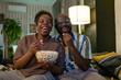 © pressmaster - Happy African couple eating popcorn during watching the comedy and laughing sitting on sofa in the living room