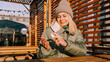 © brillianata - A young woman in a warm jacket treats her hands with an antiseptic outside. She is located in a stylish urban space with wooden decor.