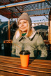 © brillianata - Happy woman on a winter sunny day at a street food fair in space for relaxing and meeting friends. She drinks coffee or mulled wine from an orange cup