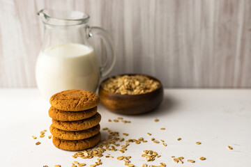 Heap oatmeal cookies and oat cereal flakes bowl on white table on milk glass jug background